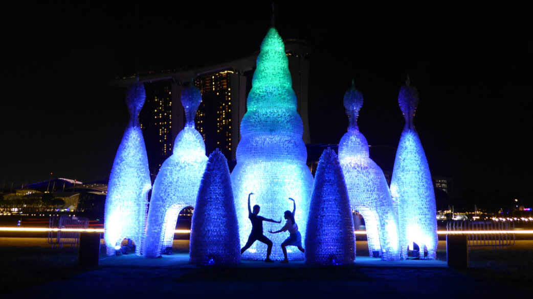 Illuminated sculpture of five blue-lit bottle towers with a taller green-tipped central spire, made from 25,000 plastic bottles. Two dancers pose in silhouette beneath, with Singapore's Marina Bay Sands visible behind.