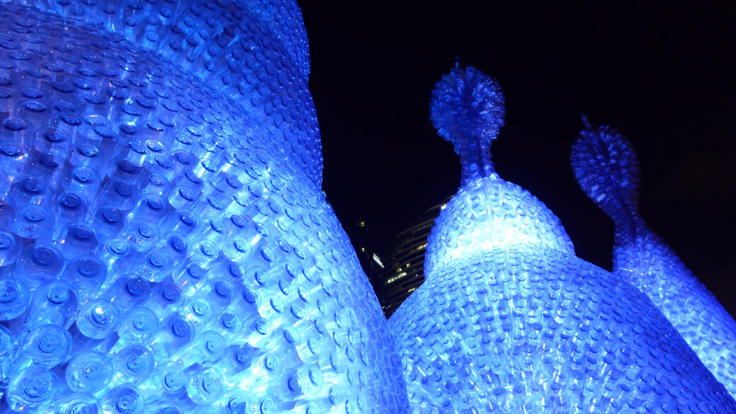 Illuminated blue sculpture resembling radiolarian sea creatures, constructed from thousands of recycled plastic water bottles, photographed at night with city buildings visible in background.