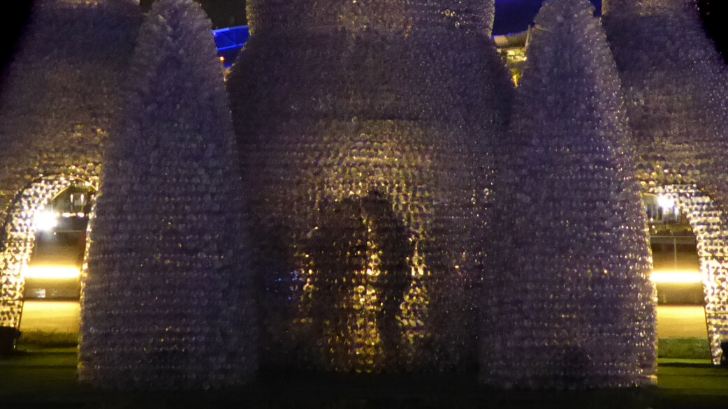 A castle-like pavilion structure made from thousands of translucent plastic bottles, illuminated from within at night, featuring turrets and arched doorways inspired by radiolarian forms.