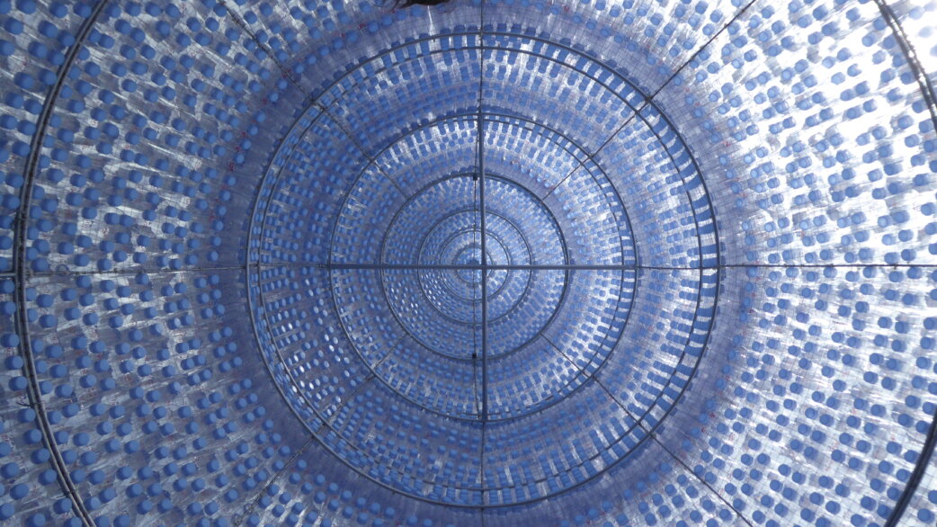Interior view looking up through a domed pavilion constructed from thousands of blue-capped plastic water bottles arranged in concentric circles, creating a translucent radiolarian-inspired structure.