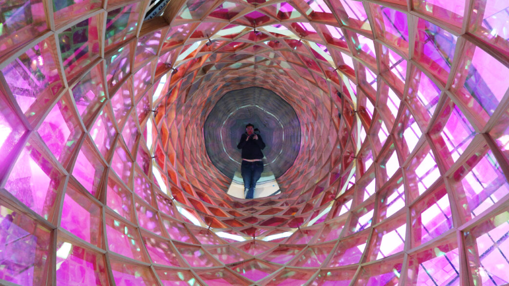 A person stands inside a cylindrical tower structure made of wooden lattice and translucent pink, orange and clear panels, with mirrored surfaces creating an infinite tunnel effect above and below.