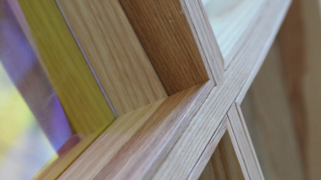Close-up detail of wooden frame joinery showing precisely mitred ash timber sections meeting at angles, with glimpses of coloured translucent panels visible through the gaps.