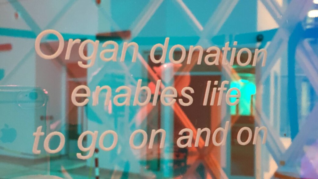 Text reading "Organ donation enables life to go on and on" printed on translucent coloured panels, with turquoise and orange light filtering through a geometric structure.