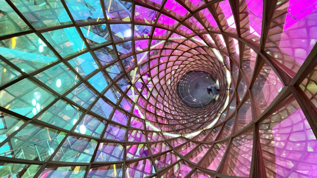 Interior view looking up through a cylindrical tower structure made of geometric coloured glass panels in turquoise, purple and pink, with a mirrored ceiling creating an infinite tunnel effect.