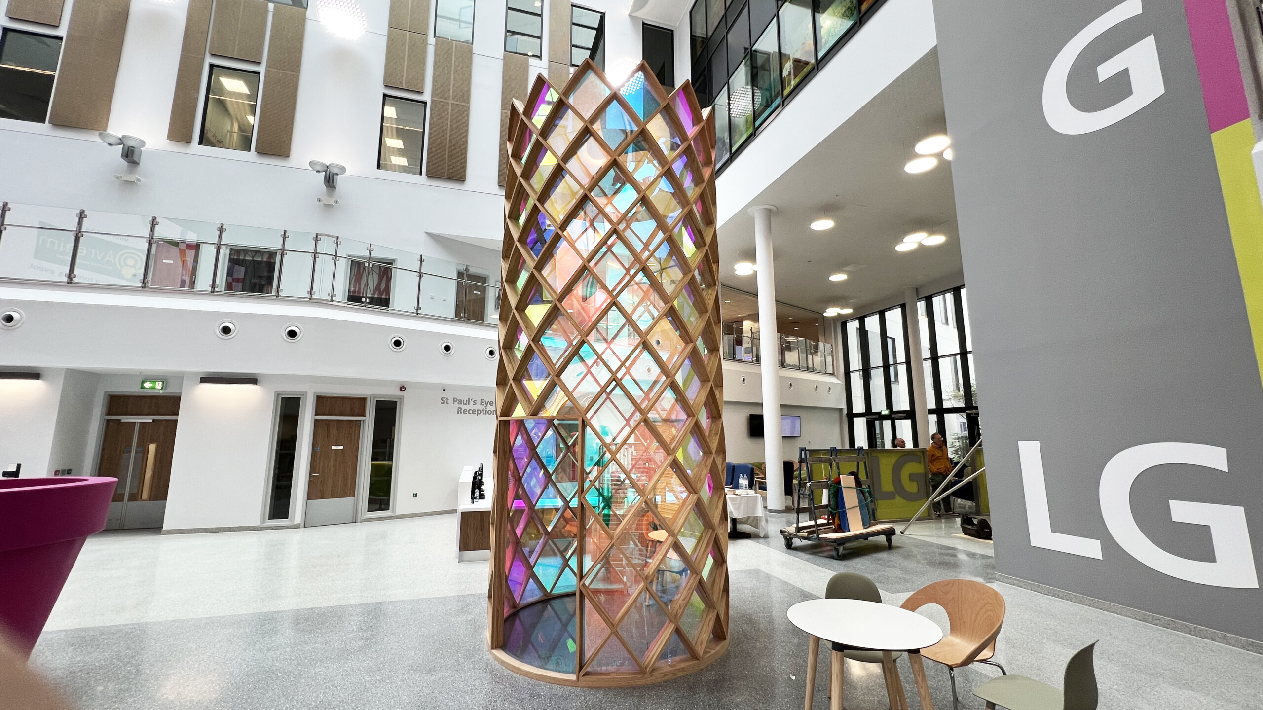 A cylindrical tower sculpture approximately 4 metres tall with a wooden lattice framework filled with iridescent coloured glass panels, situated in a hospital atrium.