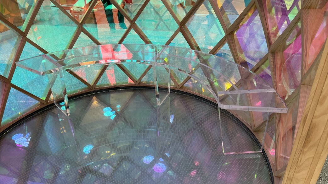 Interior view of Tribute, a cylindrical tower structure with wooden lattice framework and translucent coloured panels in blues, greens and pinks, with a circular reflective floor below.