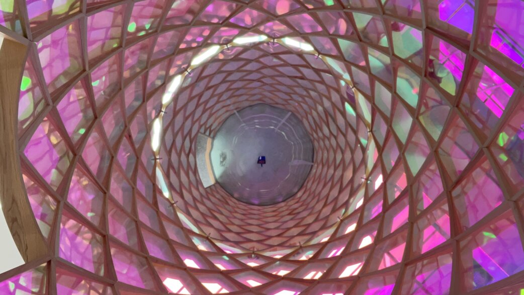 View looking up through a cylindrical tower structure with wooden lattice framework and translucent pink, purple and green panels, creating kaleidoscopic patterns with a mirrored ceiling at the centre.