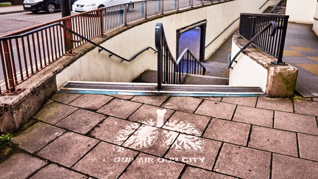 A stencilled image of human lungs with the text "Our Air Our City" cleaned onto grey paving slabs near a pedestrian underpass, created by removing surface pollution.