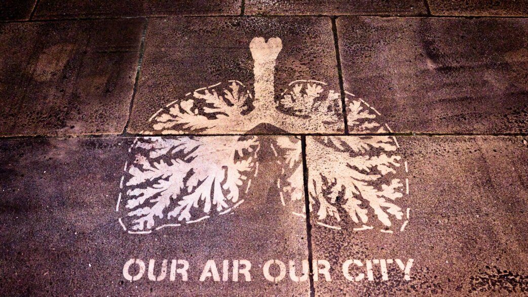 Stencilled image of human lungs made from tree-like branching patterns, created by cleaning pollution off dark paving stones, with text reading "OUR AIR OUR CITY" below.