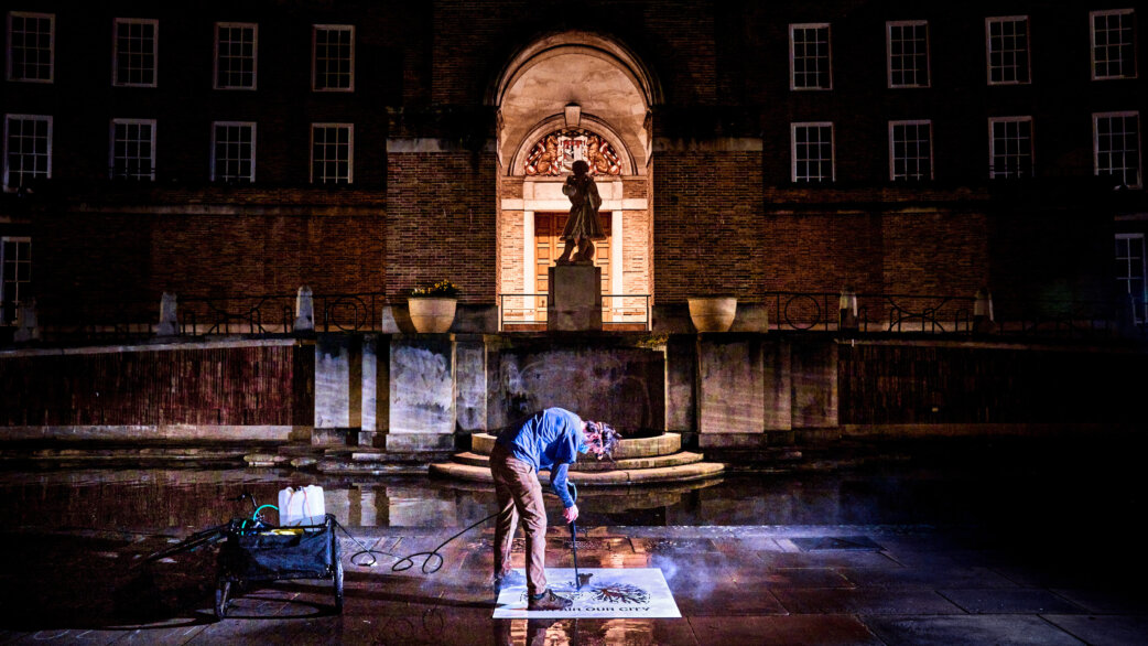 A person uses a high-pressure hose with a lung-shaped stencil to clean pollution from paving stones at night, creating clean graffiti outside Bristol City Hall.
