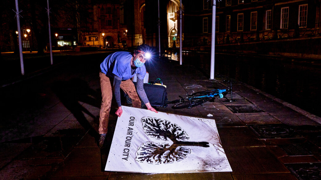 A man wearing a headlamp and face mask lifts a large stencil revealing a clean graffiti image of lungs with text "Our Air Our City" on a dirty pavement at night, bicycle nearby.
