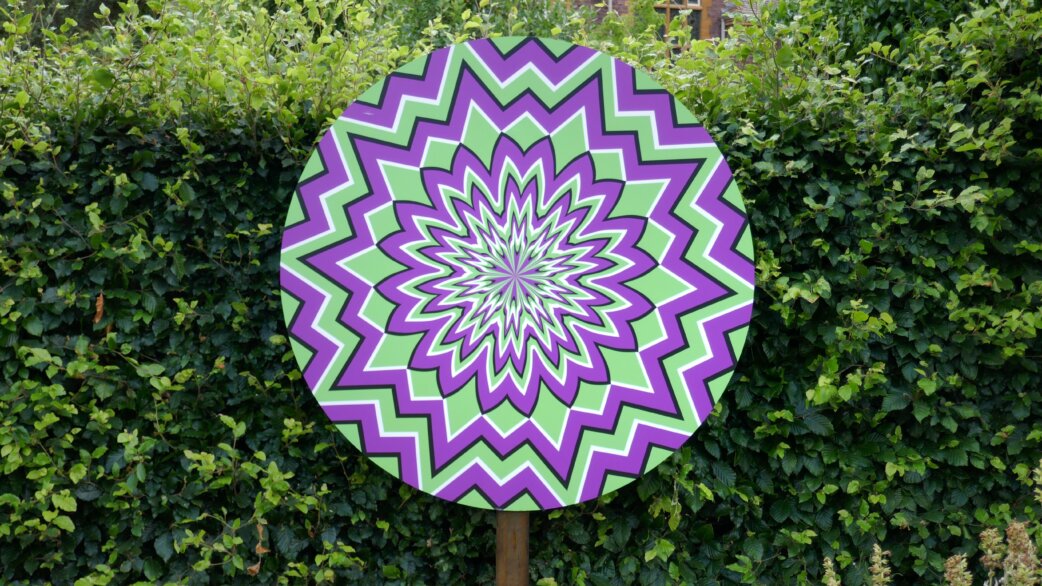 A circular sign displaying a green and purple zigzag optical illusion pattern radiating from the centre, mounted on a post against a dense green hedge.