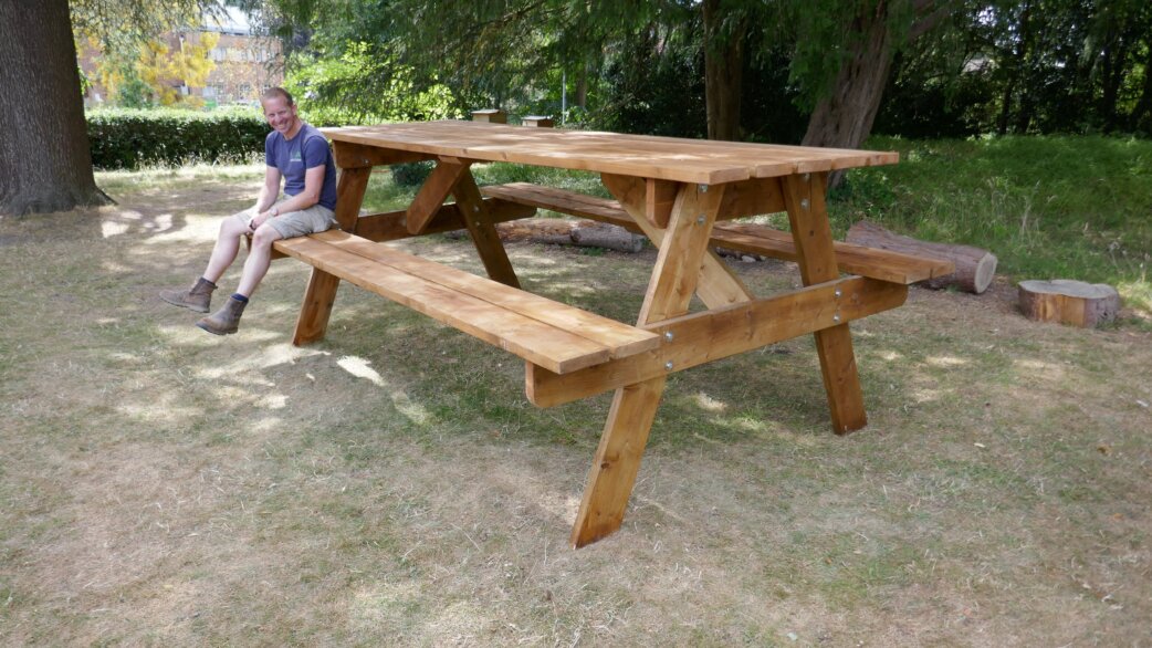 A man sits on an oversized wooden picnic table approximately twice the normal size, set in a garden with trees. The table demonstrates scale distortion as part of an optical illusion artwork.