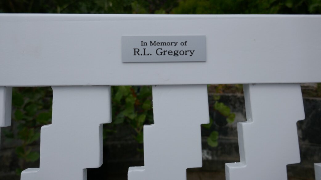 A white painted wooden bench with a small silver memorial plaque reading "In Memory of R.L. Gregory" mounted on the top rail, with foliage visible behind.