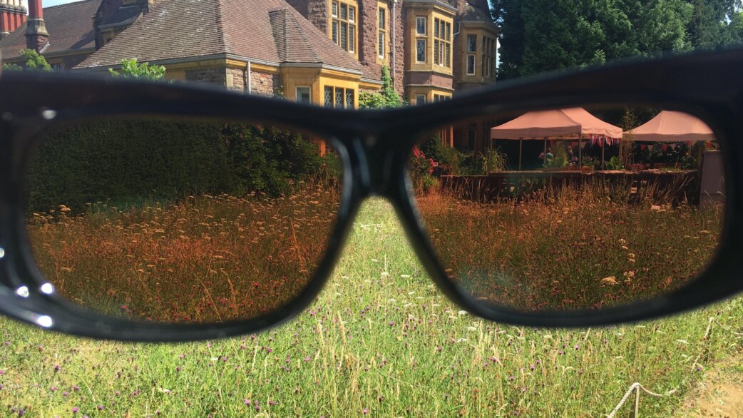 A pair of sunglasses held up against a meadow and historic stone building, demonstrating how tinted lenses alter colour perception of grass and wildflowers.