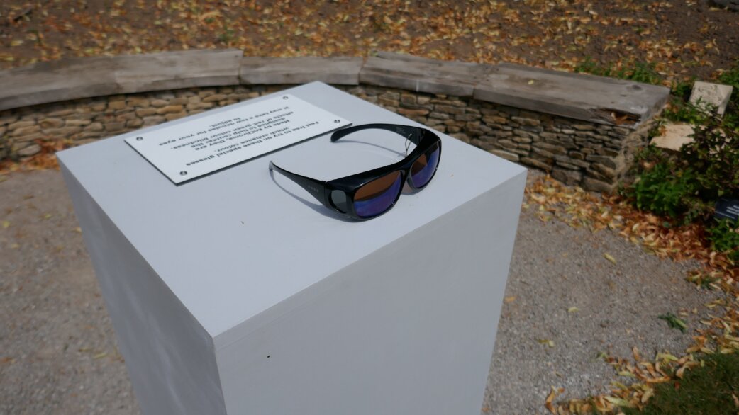 A pair of dark sunglasses with blue-tinted lenses rests on a white plinth with an information plaque, set in an outdoor garden with stone walls and autumn leaves.