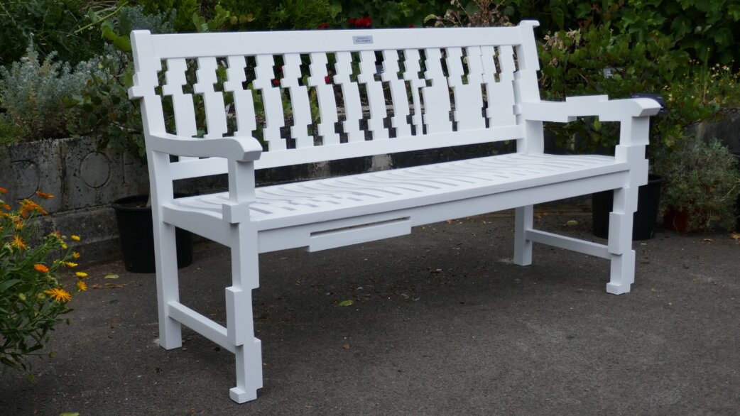 A white-painted wooden garden bench with decorative slatted backrest, positioned on gravel in front of green shrubs and potted plants at the University of Bristol Botanic Garden.