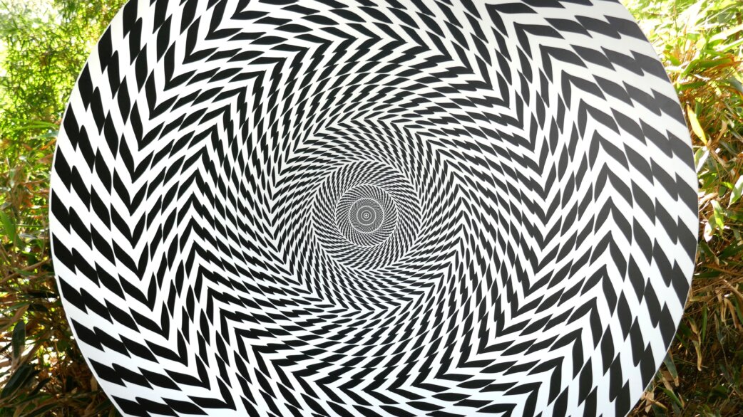 A circular black and white optical illusion sculpture featuring zigzag patterns spiralling inward to create a hypnotic vortex effect, displayed outdoors among green foliage.