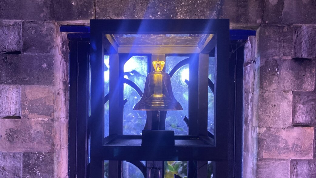 A bronze bell with golden crown displayed in a dark metal frame with decorative cutouts, illuminated by blue and purple lighting, set within a stone archway.