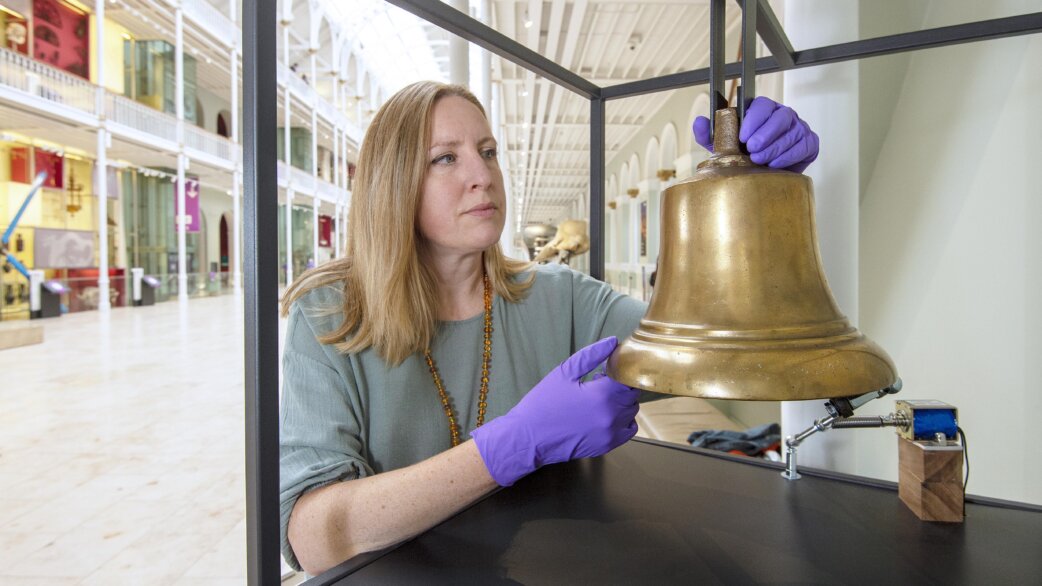 A woman wearing purple conservation gloves carefully positions a brass bell, approximately 30cm tall, in a museum display case within a grand Victorian gallery space.