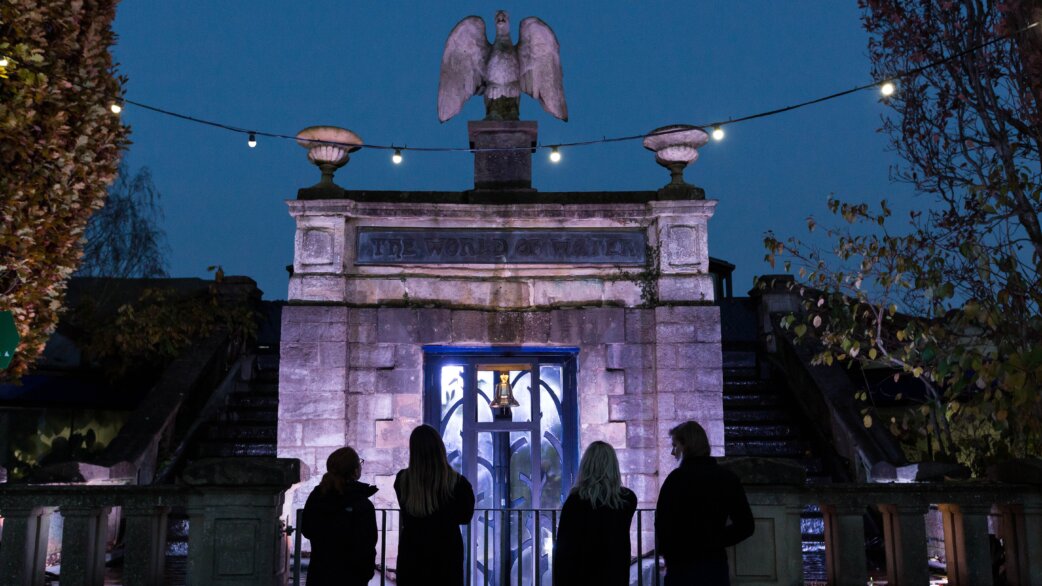 Silhouettes of four people viewing the Extinction Bell installation at dusk, housed in a stone gateway topped with an eagle sculpture and decorative urns, illuminated by string lights.