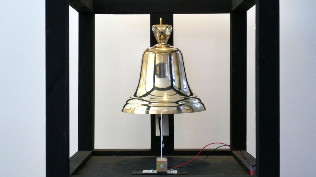 A polished brass bell mounted within a black metal frame, with an electronic striking mechanism and wires visible at its base, displayed against a white background.