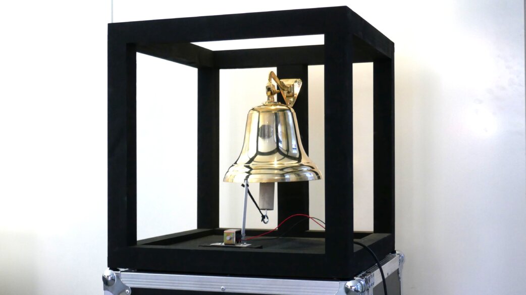 A polished brass bell mounted within a black cubic wooden frame, sitting on a flight case. Wires connect to an automated striking mechanism beneath the bell.