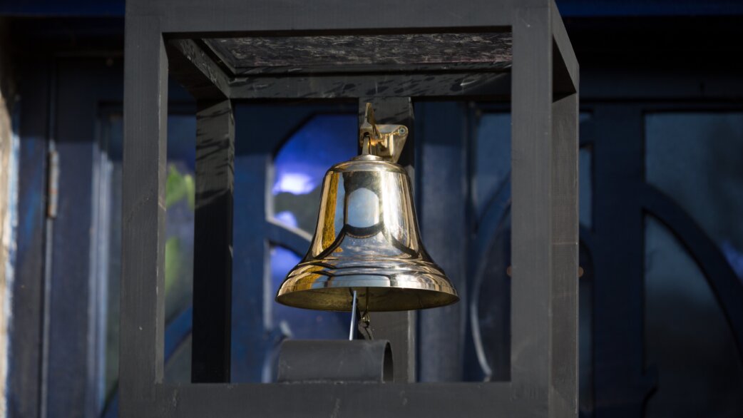 A polished brass bell housed within a dark wooden frame, mounted outdoors. The bell gleams in natural light against a deep blue background.