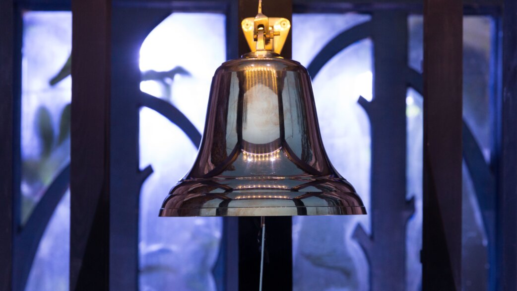 A highly polished brass bell with a golden mounting bracket, displayed in a wooden frame against blue-lit decorative glass panels with organic curved patterns.
