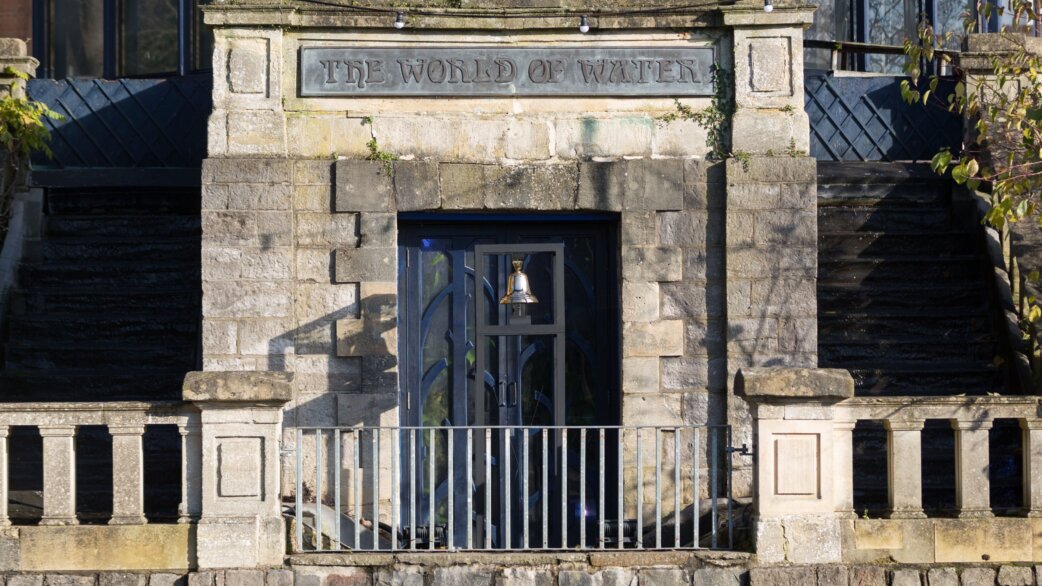 A brass bell mounted on dark metal double doors set within a grand stone entrance carved with "The World of Water" above, with balustrade in foreground.