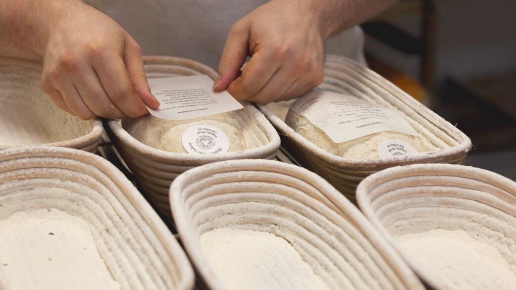 Hands placing a small printed rice paper poem onto bread dough in an oval proofing basket, with several similar baskets containing dough visible alongside.
