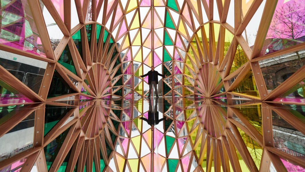 A person stands with arms outstretched inside Palm Temple, a cedar wood pavilion with dichroic coloured panels and mirrored floor, creating kaleidoscopic reflections in pink, green and yellow.