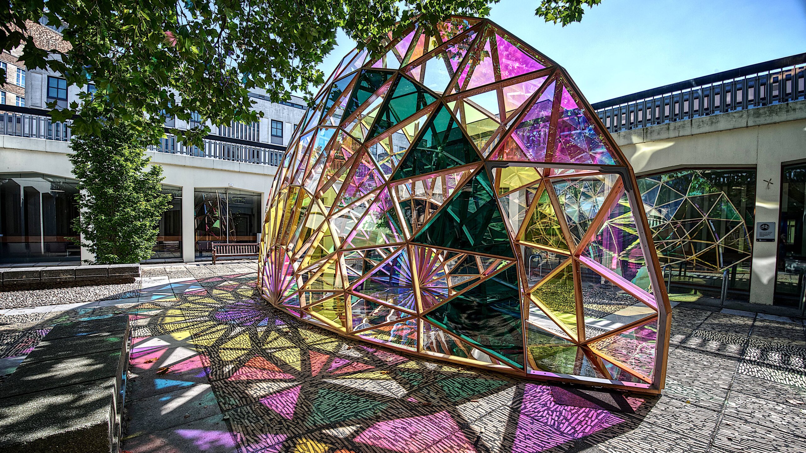 A geodesic pavilion sculpture made from cedar wood and colourful dichroic glass panels, casting vibrant multicoloured shadows on cobblestones in a university courtyard.