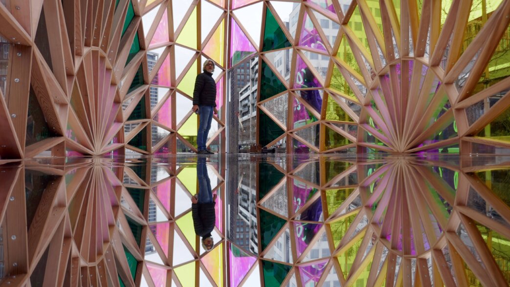 A person stands inside Palm Temple, a cedar wood pavilion with geometric radiating beams and colourful dichroic glass panels, reflected perfectly in the mirrored floor below.