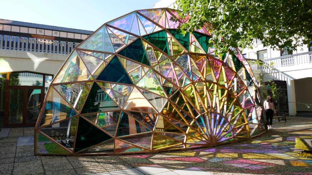 A geodesic dome pavilion made from cedar wood and colourful dichroic glass panels, casting rainbow-coloured shadows on the ground. Approximately 4 metres tall, installed in an urban courtyard.