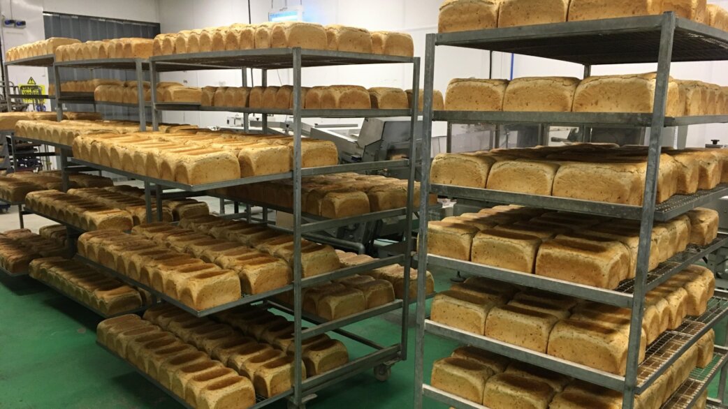 Rows of freshly baked white bread loaves cooling on metal industrial shelving racks in a commercial bakery, with a green floor visible beneath.