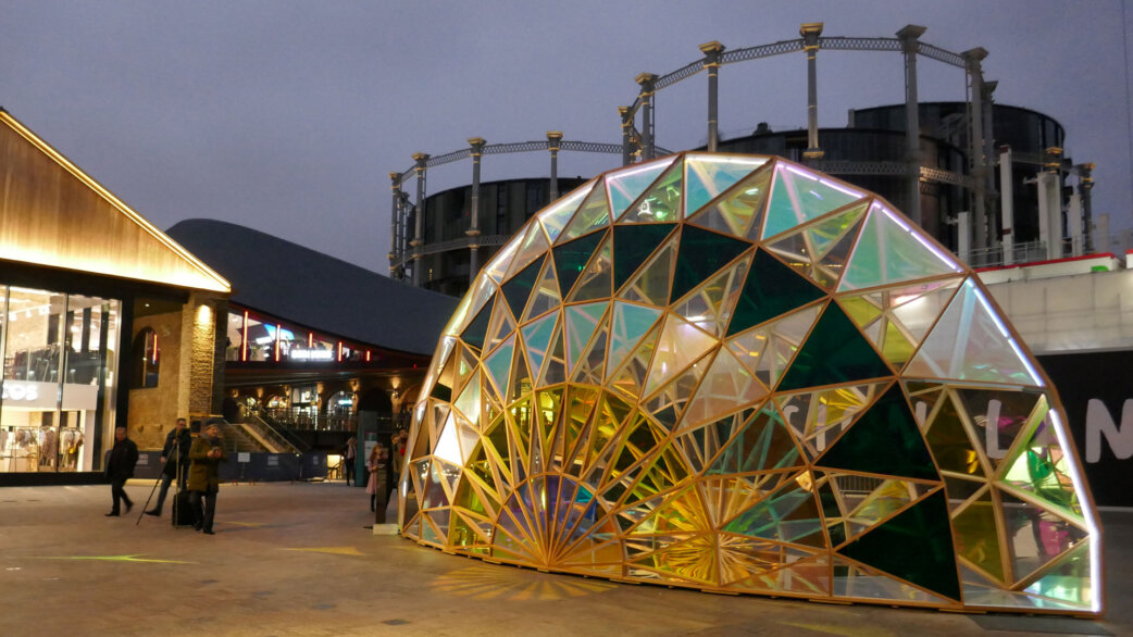 A geodesic dome sculpture made of dichroic glass panels in iridescent colours, approximately 4 metres tall, installed in an urban plaza at dusk with historic gas holders visible behind.