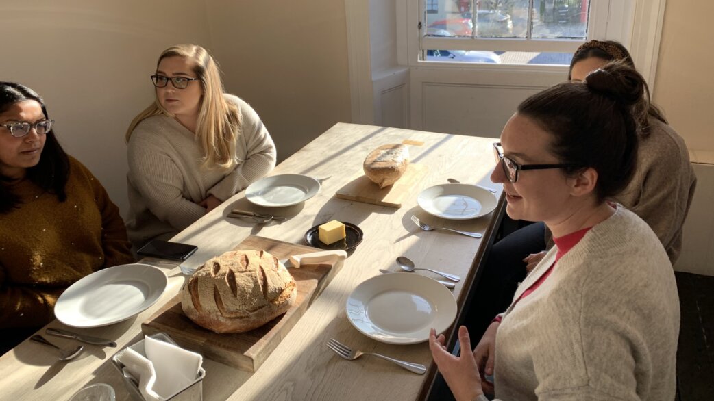 Five women seated around a light wooden table with artisan bread loaves on cutting boards, white plates, butter and cutlery, appearing to share a bread poetry tasting session.
