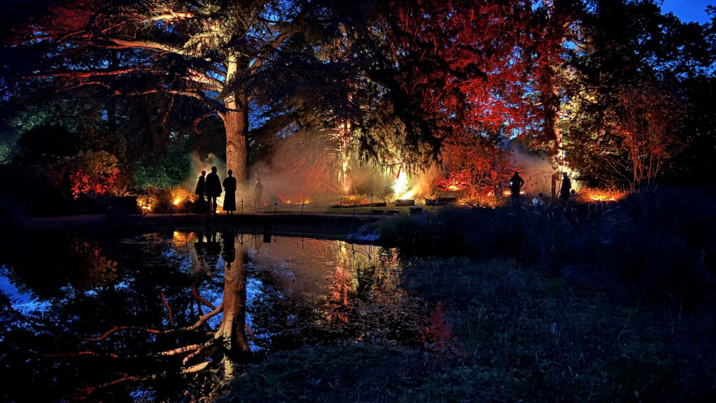Silhouetted figures standing by a reflective pond surrounded by trees illuminated in red and orange light with atmospheric smoke.