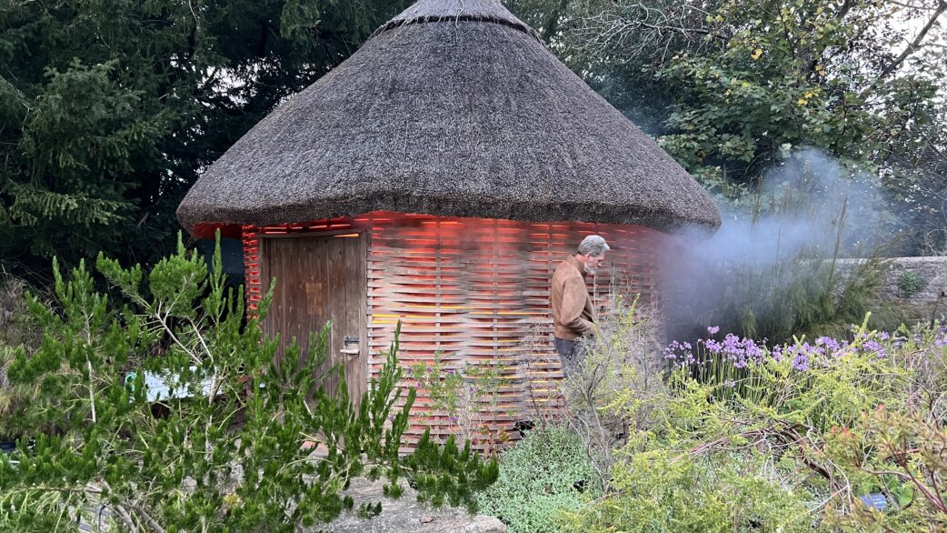 A circular thatched-roof structure with horizontal wooden slats glowing orange-red from within, smoke billowing from one side, surrounded by garden plants. A person stands nearby observing.
