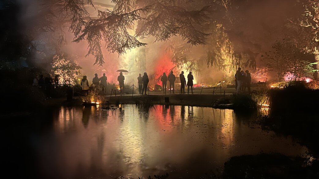 Silhouetted figures stand on a bridge over a reflective pond at night, surrounded by dramatic orange and red lighting with smoke creating a simulated forest fire effect.