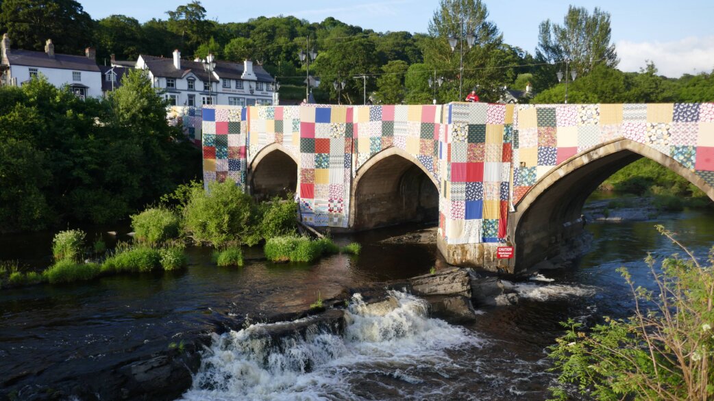 A medieval stone bridge with three arches spanning a river, wrapped entirely in colourful patchwork fabric squares. Welsh hillside houses and trees visible behind, water flowing beneath.
