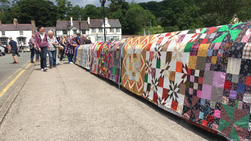 A medieval stone bridge in Llangollen wrapped in colourful patchwork quilts, approximately 60 metres long, with visitors walking alongside admiring the fabric patterns on a sunny day.