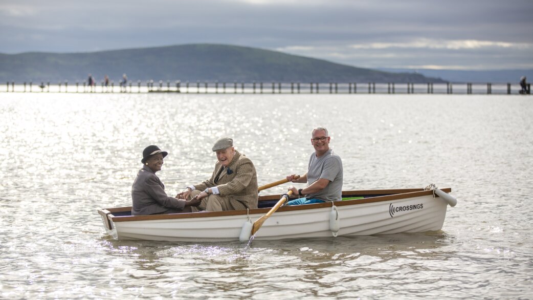 Three people in a white rowing boat named Crossings on calm water, with a pier and hills in the background. One person rows while two elderly passengers smile.