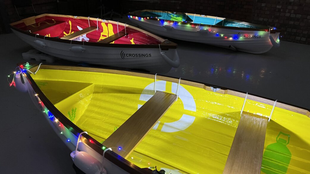 Three colourful rowing boats decorated with fairy lights floating on dark water. Each features painted designs and "Crossings" branding, with wooden seats and rope details.