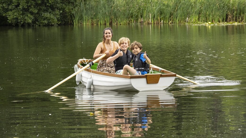 A woman rows a white wooden boat on a calm green lake while two children in life jackets sit at the front giving thumbs up, with reeds in the background.