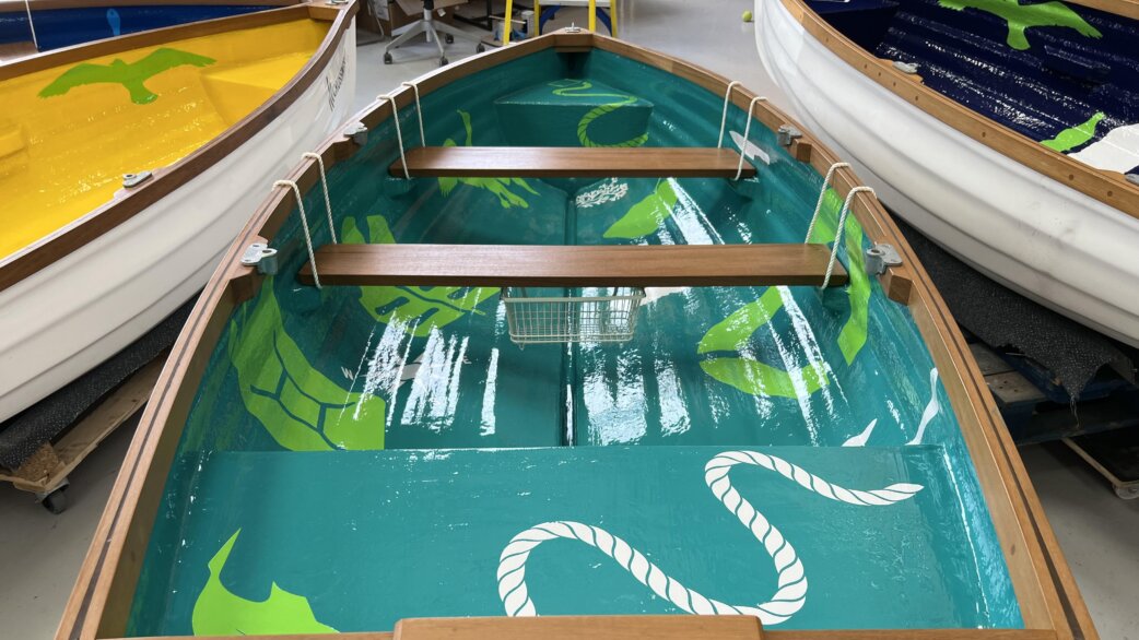Interior view of decorated rowing boats with glossy painted designs featuring green sea turtles, birds and rope motifs on turquoise backgrounds, with wooden thwarts and rope handles.