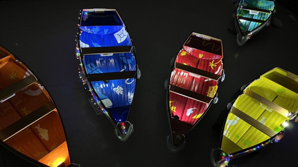 Aerial view of illuminated rowing boats floating on dark water at night, decorated in vibrant blue, red, yellow and orange with painted marine imagery and string lights.