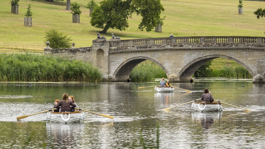 Three white rowing boats on a calm lake, each carrying visitors listening to audio stories. A historic stone arched bridge and green parkland visible behind.