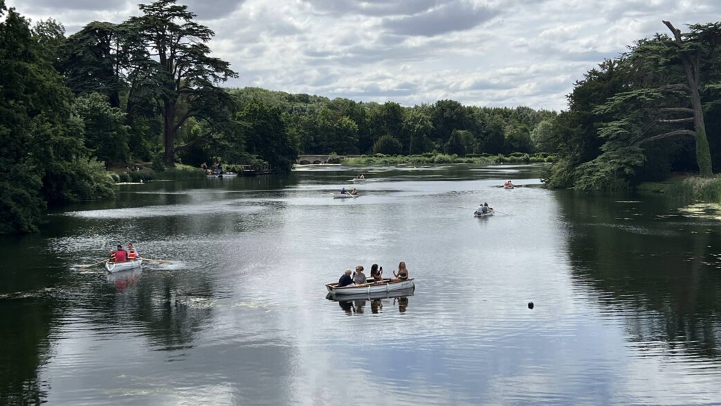 Several rowing boats carrying visitors float on a calm lake surrounded by mature trees under a cloudy sky, part of Luke Jerram's Crossings audio installation at Compton Verney.
